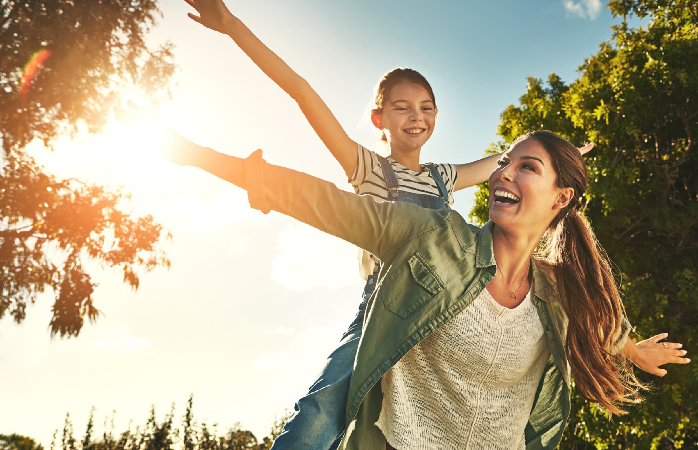 Mother giving her young daughter a piggyback outdoors, both laughing with arms outstretched in sunshine near trees.