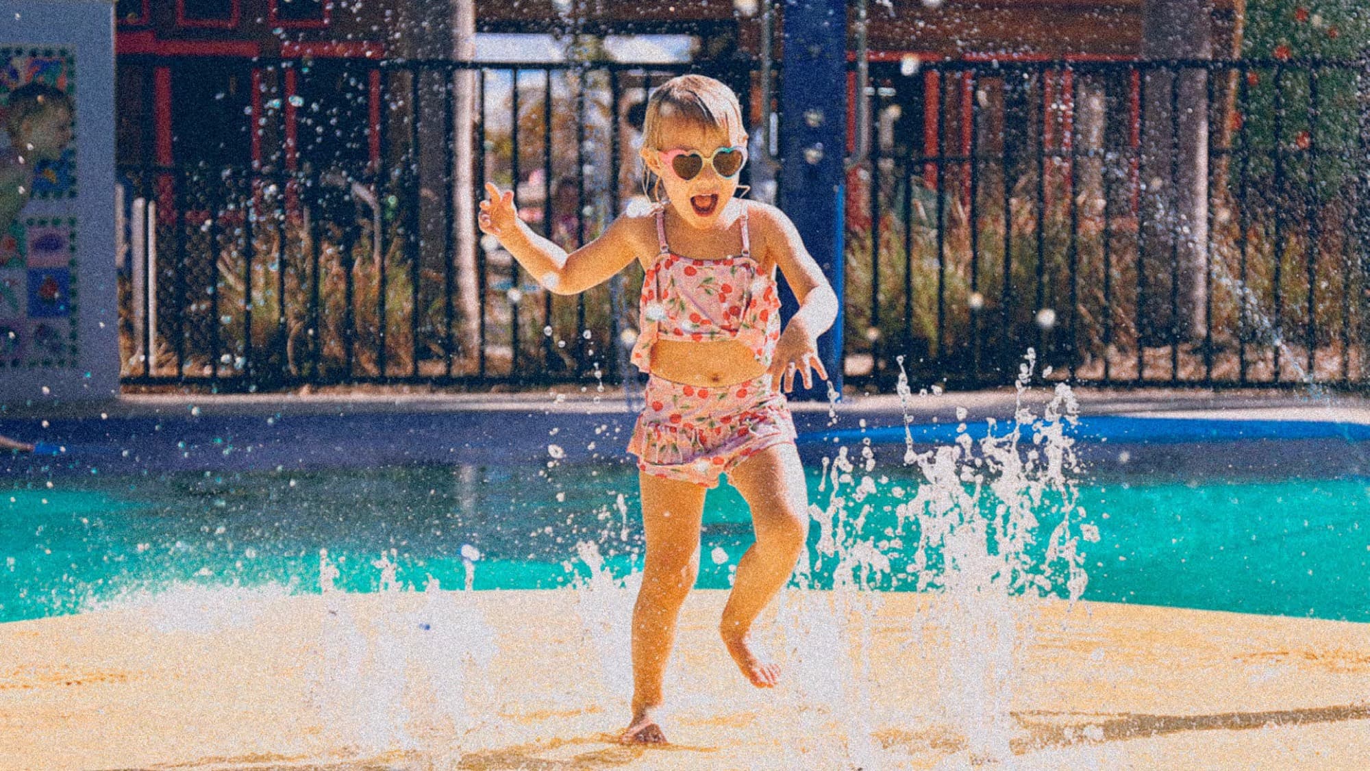 Young girl in heart-shaped sunglasses splashing through a water play area at Joy, Arabian Ranches 3, Dubai.
